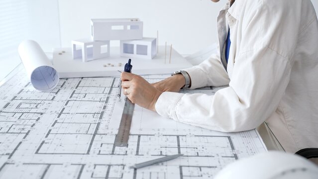 Professional architect's hands study detailed blueprints and building plans on a drafting table, with a 3d house model nearby, focusing on residential design and engineering decisions