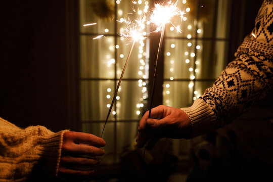 Couple Hands Holding Burning Sparklers in Warm Cozy Sweaters with Golden Christmas Lights Bokeh Background on New Year Eve. Happy new year, atmospheric eve