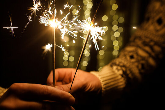 Happy New Year. Close Up of Hands Holding Glowing Bengal Sparklers on Warm Golden Bokeh Lights Background. Magic moments, New Year celebration, holiday cheer.