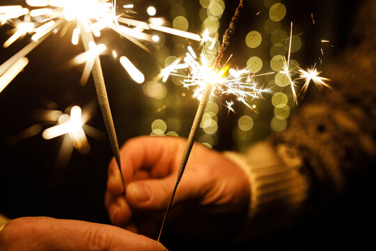 Macro shot of hands holding bright sparkling bengal lights with golden bokeh lights background. Magic moments, New Year celebration, warmth and holiday cheer.
