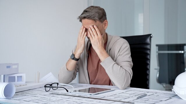 Stressed architect feeling frustrated and overwhelmed at his office desk. Covering his face with hand while working on architectural project blueprints and building models