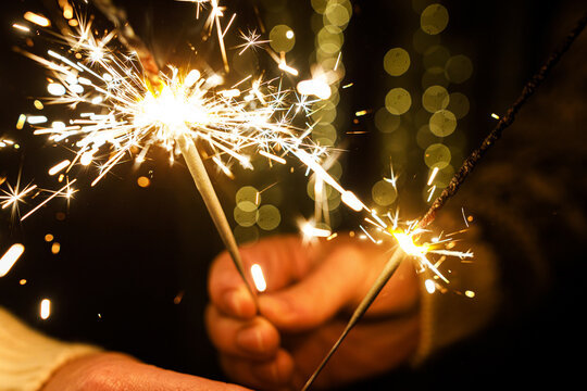 Macro View of Brightly Burning Sparkler Held by Hands with Golden Bokeh Garland Background. Togetherness and New Year celebration.