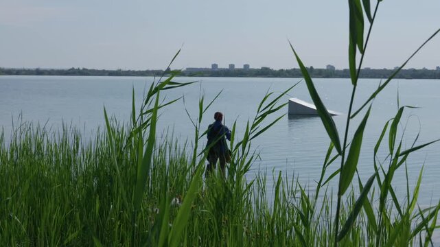 A fisherman stands near the edge of a calm lake surrounded by tall green reeds. In the background, a wakeboard ramp floats on the open water under daylight. The sky remains clear.