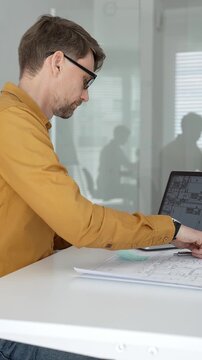 Engineer working on architectural blueprints and plans, typing on a laptop with a compass, ruler, safety helmet, glasses, and a building model on the desk