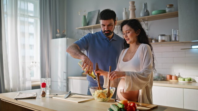 Married couple cooking salad kitchen. Husband adds oil pregnant wife mixing.