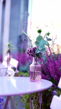 Empty lavender coffee table arranged outdoors on city street with glass vase filled with fresh purple flowers. Minimal table setting creates inviting urban cafe atmosphere while soft natural daylight