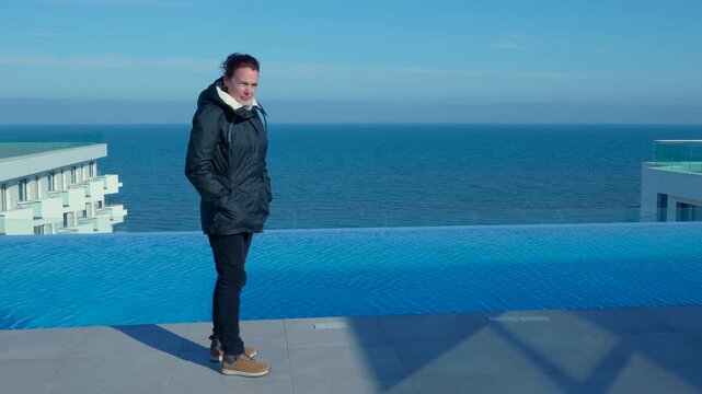Woman standing by infinity pool enjoying the ocean view. Young woman standing on a rooftop with an infinity pool, contemplating the serene ocean view from a luxury hotel on a sunny day