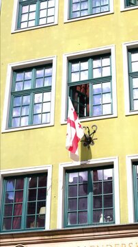 Georgian national flag mounted on building facade in urban setting. White fabric with red cross design hangs prominently against architectural exterior, clearly marking cultural diplomatic or