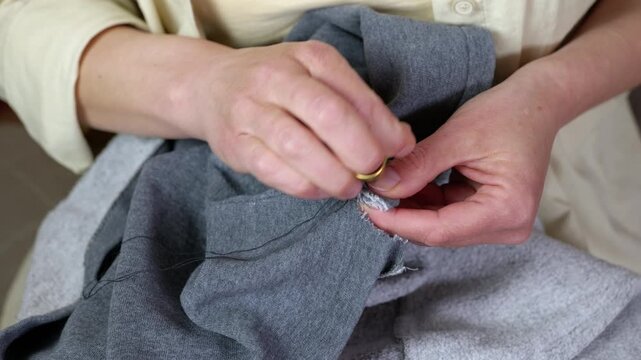 Woman mending and hand sewing gray fabric with a needle. Woman's hands using a needle and thread to manually repair a hole in a gray garment, demonstrating a sustainable lifestyle