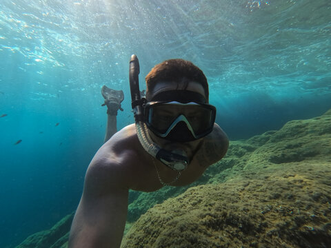 Man snorkeling underwater in mask and fins near rocky seabed in clear blue sea with sun rays breaking surface