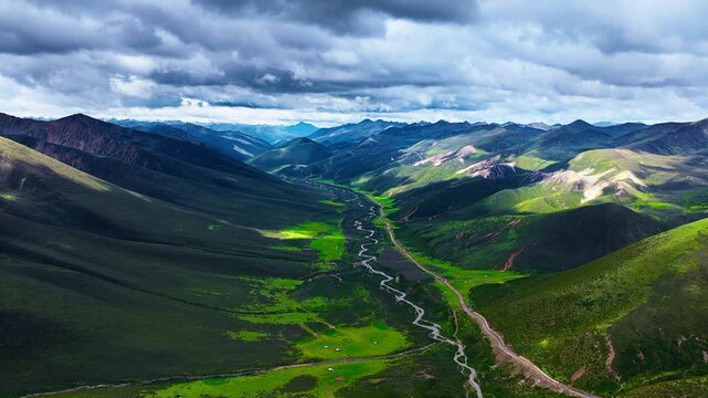 Aerial view of a sprawling green mountain valley with a winding stream and dirt road under dramatic cloudy daylight.