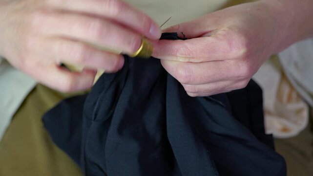 Close up of woman's hands sewing fabric. Woman's hands carefully sewing black fabric with a needle and thread, using a golden thimble to push the needle through the cloth