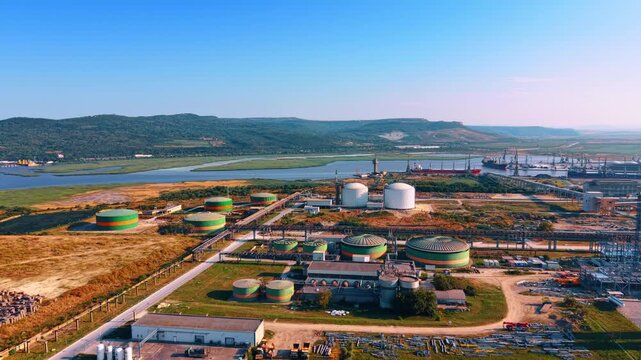 Diverse cylindrical storage towers connected by the pipelines. Port on the river and green hills in the backdrop.