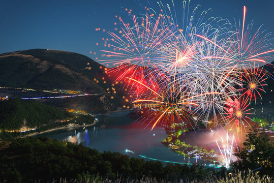 Aerial view of vibrant fireworks illuminating the night sky over the calm waters of Lago di Fiastra surrounded by dark mountains in Fiastra, Marche, Italy.