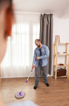 Smiling man uses a mop on the wooden living room floor while another person appears softly blurred in the foreground during everyday housework and floor care.