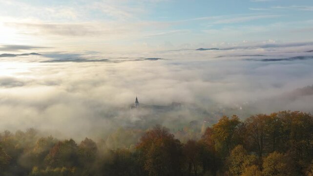 AERIAL: Sea of morning fog blankets the valley around Sticna Abbey, leaving its church tower and rooftops visible through the mist. Autumn woodland adds warm colour to the tranquil hilly landscape.