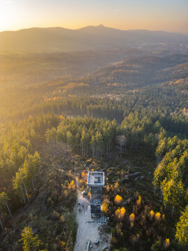 Aerial view of the Cisaruv kamen lookout tower surrounded by a dense forest during the golden hour, with distant mountains under a warm sunset sky in Liberec 30, Liberec Region, Czechia.