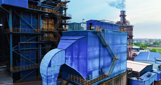 Industrial factory complex featuring blue metal structures, external ductwork, catwalks, and a smoking chimney under overcast daytime light.