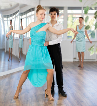 Couple of positive interested teenage dancers, boy and girl preparing for performance, rehearsing ballroom dance in spacious classroom while female teacher observing from background..