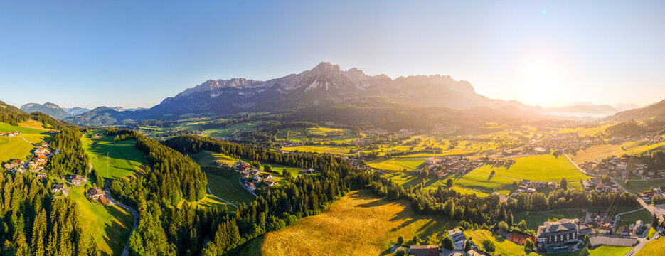 Aerial view of the Wilder Kaiser mountain range rising above lush green alpine meadows, dense forests, and a village bathed in warm golden hour sunlight in Wilder Kaiser, Tirol, Austria.