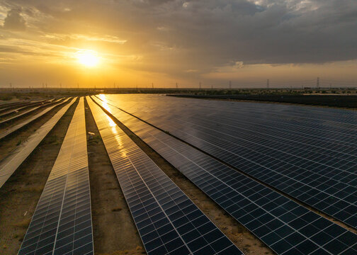 Aerial view of a vast solar farm with rows of photovoltaic panels reflecting the golden sunset light under a dramatic sky in Rajasthan, India.