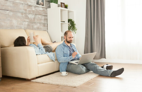 Man uses a laptop from the carpet while a woman reads on the sofa behind him, emphasizing relaxation, leisure, and downtime in daily home life.