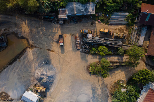 Aerial view of a construction site featuring heavy machinery, piles of gravel, and a truck parked on a dirt lot surrounded by lush greenery in Cilongok, Central Java, Indonesia.