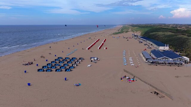 Wide horizontal aerial drone video of Katwijk aan Zee sandy beach resort, North Sea shoreline, coastal dunes, beach cabins, and kitesurfers in South Holland, Netherlands on late afternoon