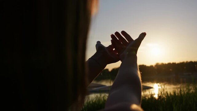 Hands stretch upward toward the setting sun over a calm lake shoreline. Bright backlighting creates a warm silhouette atmosphere among tall grass, inspiring a sense of deep peace.