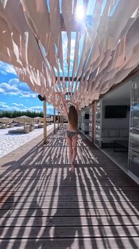 Back view of young woman in bikini dancing and walking forward along wooden terrace pathway at beach resort, slender girl enjoying summer vacation on sunny day, outdoor resort lifestyle concept