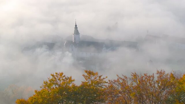 AERIAL, CLOSE UP: Historic Sticna Abbey appears almost dreamlike through dense morning mist, with its distinctive steeple rising above pale fog and warm autumn foliage in peaceful Dolenjska, Slovenia.