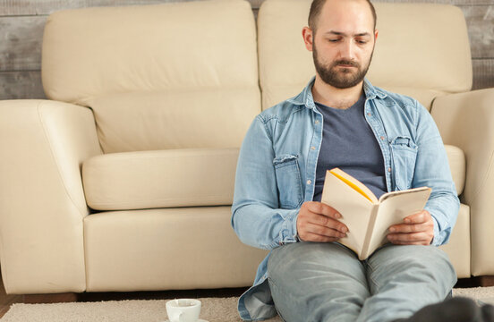 Young man reads a book beside a coffee cup at home during a quiet period of leisure, concentration and screen-free relaxation, highlighting rest.