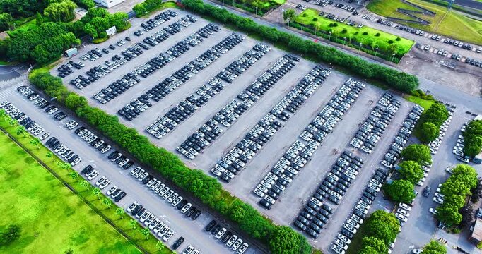 Aerial view of an outdoor parking lot filled with rows of parked cars, bordered by green lawns and trees under bright daylight.