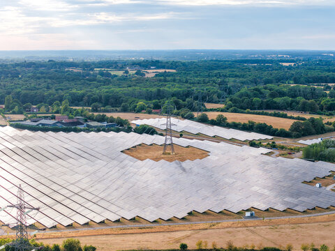 Aerial view of a large solar farm with rows of photovoltaic panels and electricity pylons set amidst green fields and trees in Hook, England, United Kingdom.