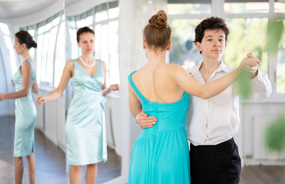 Positive motivated adolescent dance duo, boy and girl, refining slow foxtrot technique in dance hall while interested female coach providing expert guidance