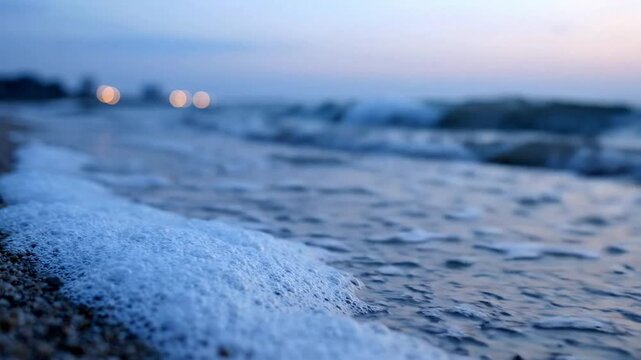 Serene ocean waves crashing on pebble beach at twilight, soft blue hour coastal background, with bokeh city lights