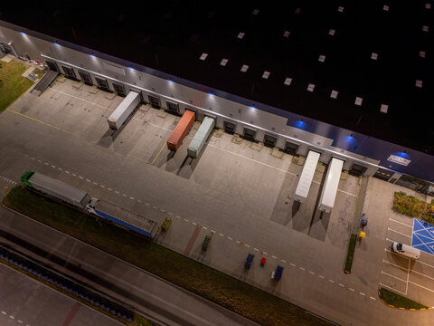 Aerial view of a logistics warehouse at night with loading docks, parked semi-truck trailers, and illuminated parking area in Gdansk, Poland.