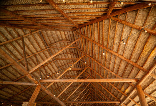 A symmetrical view of a wooden roof framework with dry thatch and hanging string lights. Traditional rustic architecture and natural design.