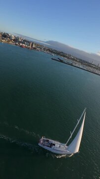 Fast low aerial flight over the sea passing close to a sailboat and approaching the Auckland skyline in New Zealand, captured as dynamic vertical footage.