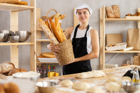 Positive teenage girl wearing pinafore holding baguettes in round wicker basket in bakery shop