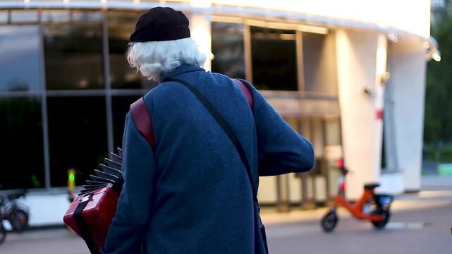 Elderly Musician Carrying Red Accordion Walking Near Modern Building
