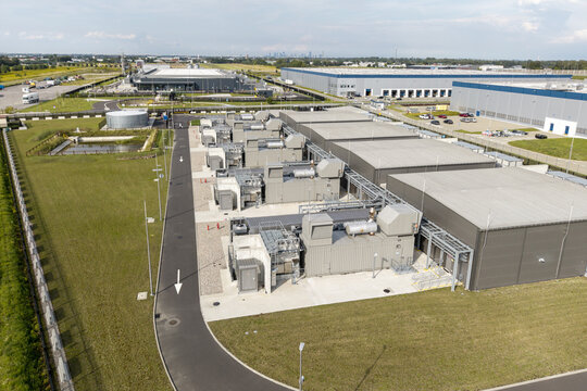 Aerial view of a modern industrial facility featuring large warehouse buildings, rooftop HVAC systems, paved access roads, and surrounding green spaces in Poland.