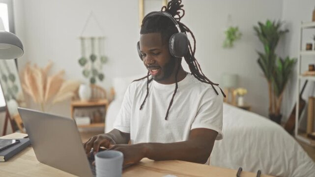 Young man typing on laptop with headphones at bedroom desk, smiling and leaning forward; remote work productivity.