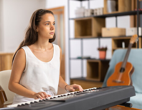 Young woman wears shorts, sits at synthesizer and plays music in room