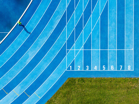 Aerial view of a person running on a blue athletic track with lanes numbered one to eight and adjacent green grass in Poland.