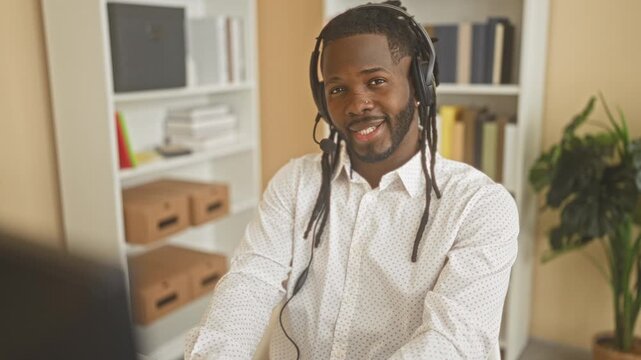 Man call center agent wearing headset and white shirt smiling at camera while seated at computer desk with bookshelf in building; friendly support.