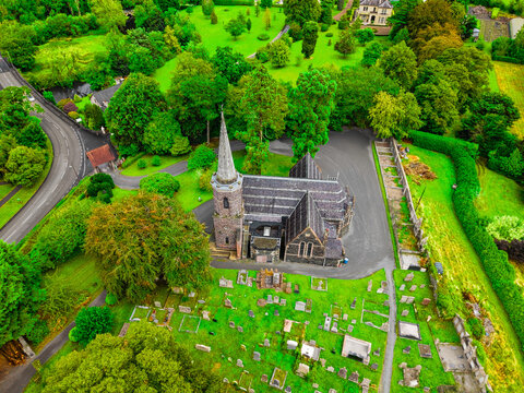 Aerial view of Drumbeg Parish Church with its stone spire surrounded by a historic graveyard and lush green trees in Drumbeg, Northern Ireland, United Kingdom.