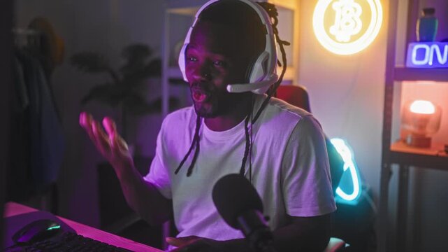 Young man with headset gestures with hands at computer keyboard and microphone while streaming games in studio; joyful focus.