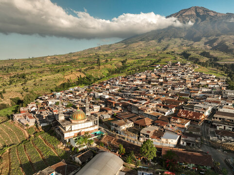 Aerial view of Kruwisan mosque nestled among dense village rooftops and terraced agricultural fields with a towering mountain in the background in Kledung, Central Java, Indonesia.