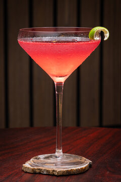 Pink cosmopolitan cocktail in martini coupe glass. Lime twist adorns the rim. Dark wood table create depth. Sleek, elegant, sophisticated mood